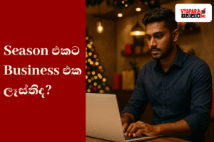 උත්සව සමය | A realistic photo of a young Sri Lankan entrepreneur working on a laptop in a warmly lit shop decorated for Christmas, with festive lights and a tree in the background. The left side features a red panel with Sinhala text reading “Season එකට Business එක ලෑස්තිද?” and the Vyapara.lk logo appears in the top right corner.