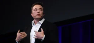 Elon Musk | A man in a black suit and white shirt stands on a stage, speaking with both hands raised near his chest. He looks slightly to the side with a focused expression, lit by soft stage lighting against a dark backdrop with blue accents.