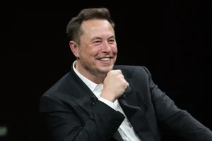 Elon Musk | Smiling middle-aged man in a black suit and white shirt, seated on a dark stage, resting his chin on his hand, appearing relaxed and engaged during a public discussion.