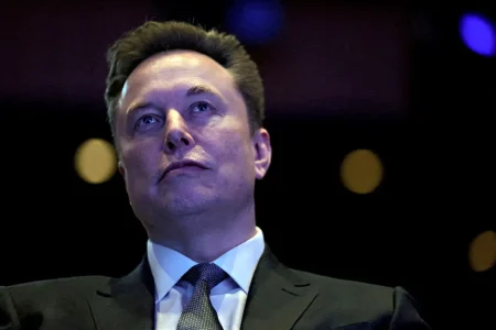 Close-up of Elon Musk in a dark suit and tie, looking upward under blue-toned stage lighting, with a blurred background of warm circular lights.