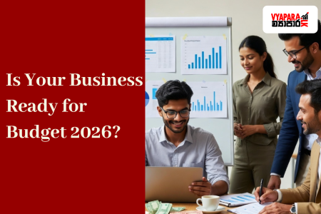 Budget | A group of young Sri Lankan entrepreneurs discussing financial charts and budget reports in a modern office. A man seated at a desk works on a laptop while others stand nearby, smiling and analysing graphs on a board. The text “Is Your Business Ready for Budget 2026?” appears on a red background beside the Vyapara.lk logo.