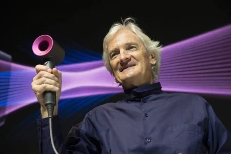 James Dyson smiling while holding a modern Dyson hair dryer, with colorful airflow graphics in the background, representing innovation and technology