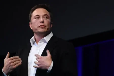 Elon Musk | A man in a black suit and white shirt stands on a stage, speaking with both hands raised near his chest. He looks slightly to the side with a focused expression, lit by soft stage lighting against a dark backdrop with blue accents.