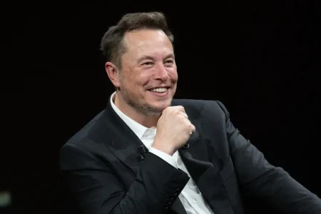 Elon Musk | Smiling middle-aged man in a black suit and white shirt, seated on a dark stage, resting his chin on his hand, appearing relaxed and engaged during a public discussion.
